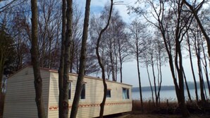 A single-story mobile home with a flat roof, positioned on a grassy area surrounded by trees. - Mobile Home im Ferienpark mit Seezugang (Dabel)