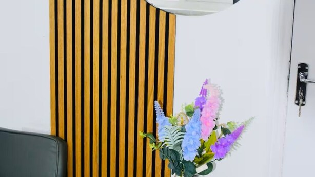 Ett rum med en vägg klädd med träribbor, en rund spegel, en vas med blommor, ett bord och en grön soffa.