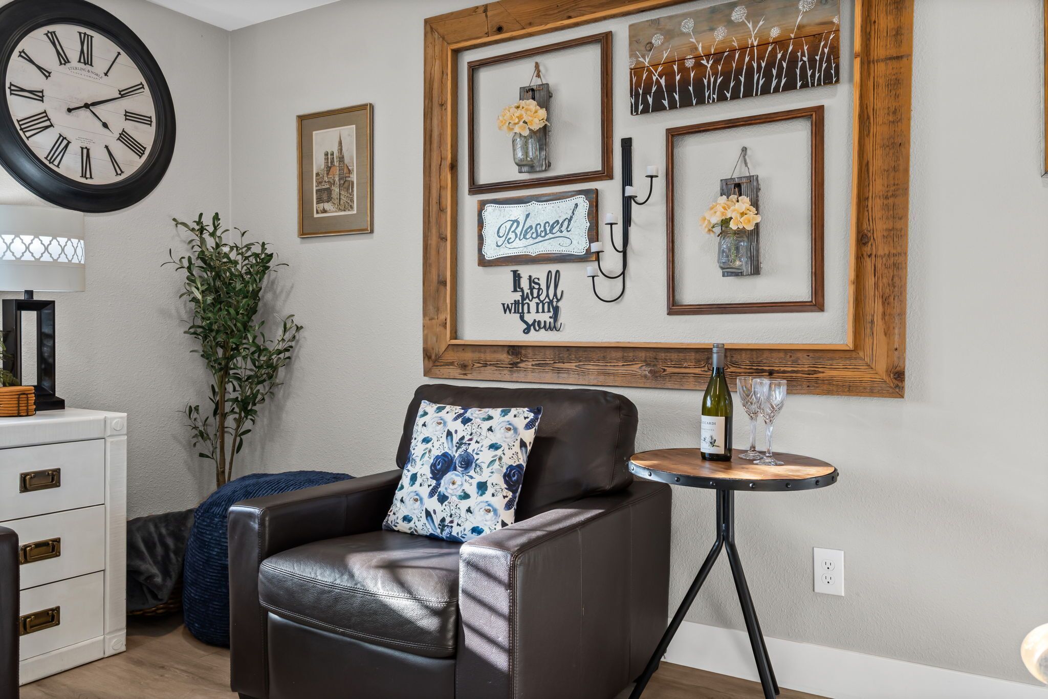 A living room with a brown leather sofa, a small round table with a bottle and glasses, a wall clock, and framed artwork.