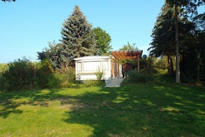 Ein kleines, weißes Gebäude mit einer Holzpergola in einer grünen Umgebung mit Bäumen.