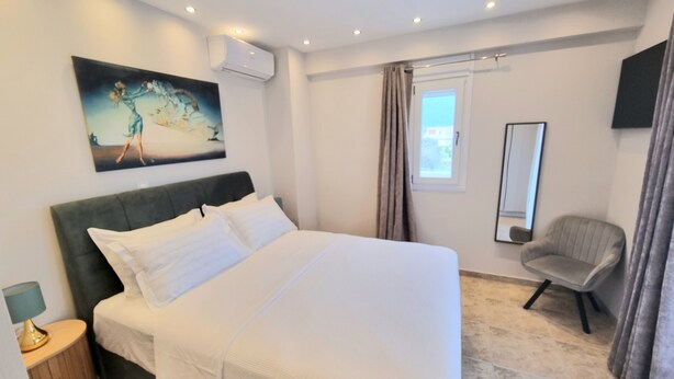 A hotel room with a large bed, a chair, a painting on the wall, and an air conditioning unit.
