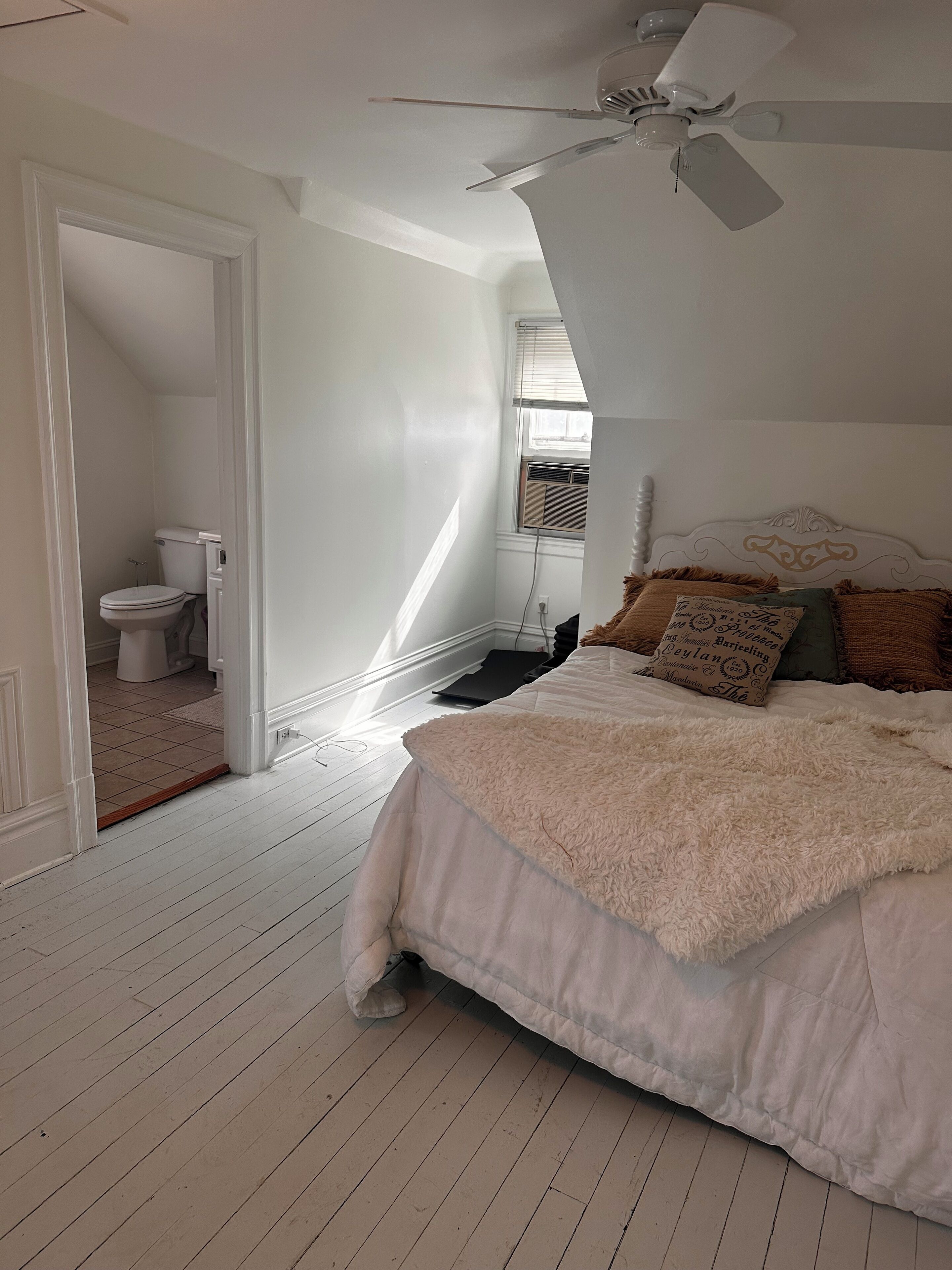 Une chambre avec un grand lit, un ventilateur de plafond, une fenêtre avec des stores et une vue sur la salle de bain.