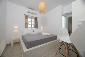 A modern bedroom with a bed, bedside tables, a chair, and a hanging light fixture. - Kasteli Luxury Rooms (Perissa)