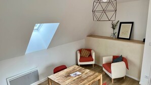 A modern apartment with a dining area, two red chairs, a wooden table, a geometric pendant light, a window, and a radiator.