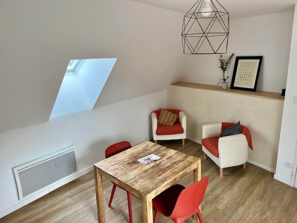 A modern apartment with a dining area, two red chairs, a wooden table, a geometric pendant light, a window, and a radiator.