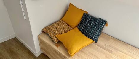 Rincón de una habitación con un banco de madera, dos almohadas y un espejo fijado a la pared.