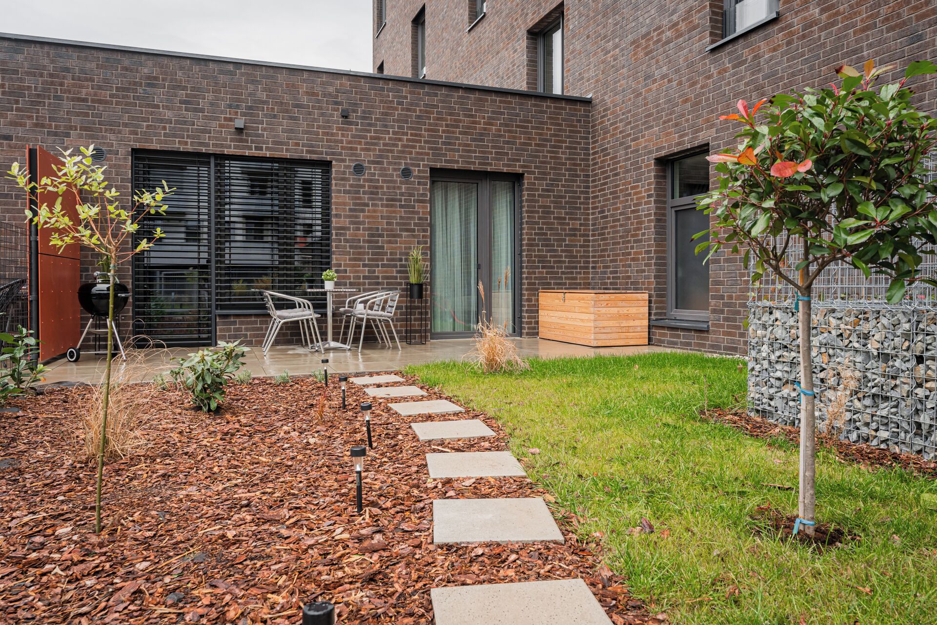 A modern brick building with a patio area featuring a grill, outdoor seating, and a garden path.