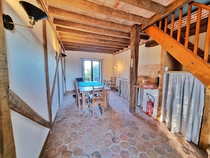 A stone-floored room with a wooden table and chairs, a staircase, and a window with curtains.
