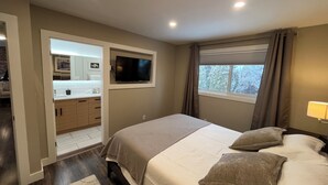 A bedroom with a bed, a TV mounted on the wall, a window with curtains, and a bathroom visible through an open door. - Close to Wineries, The Beach, trails, and Downtown (Penticton)