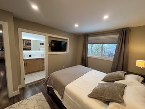 A bedroom with a bed, a TV mounted on the wall, a window with curtains, and a bathroom visible through an open door. - Close to Wineries, The Beach, trails, and Downtown (Penticton)
