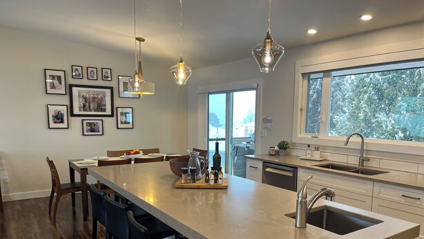 A modern kitchen with a large island, bar stools, pendant lights, and a view of the outdoors through a window. - Close to Wineries, The Beach, trails, and Downtown (Penticton)