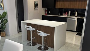 Une cuisine moderne avec des meubles de couleur sombre, un plan de travail blanc et des appareils en acier inoxydable. On y trouve un petit îlot central avec deux tabourets de bar.