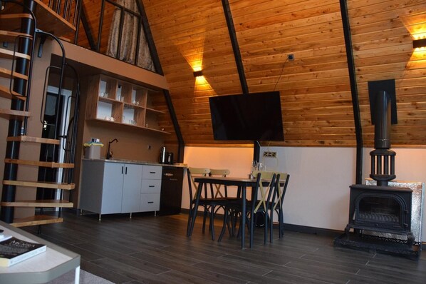Un intérieur de chalet chaleureux avec un plafond en bois, un coin repas avec une table et des chaises, une cuisine équipée de placards et d’une cuisinière, et un espace nuit en mezzanine accessible par une échelle.