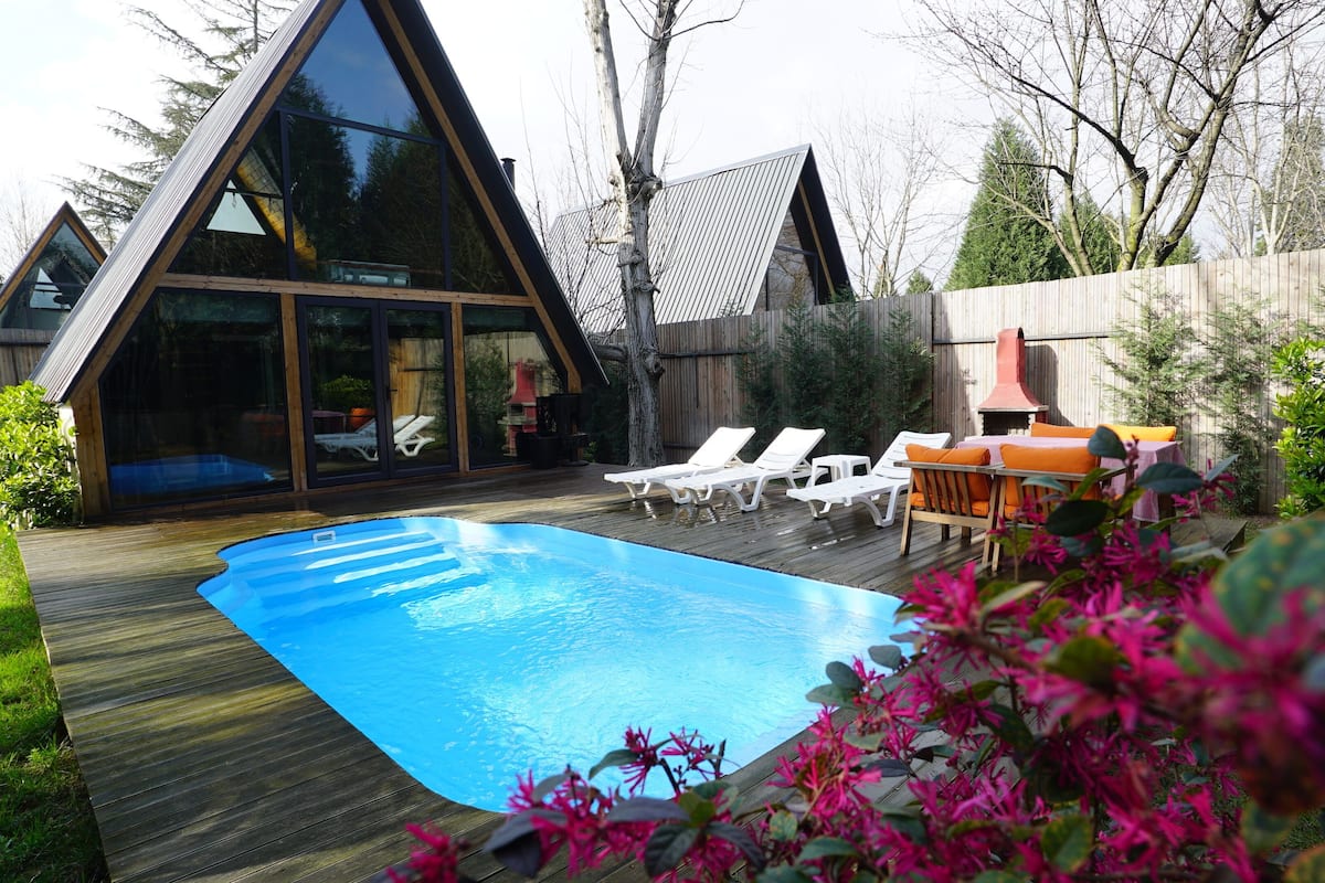 Een modern A-frame huis met een zwembad, terrasstoelen en een barbecueplek.