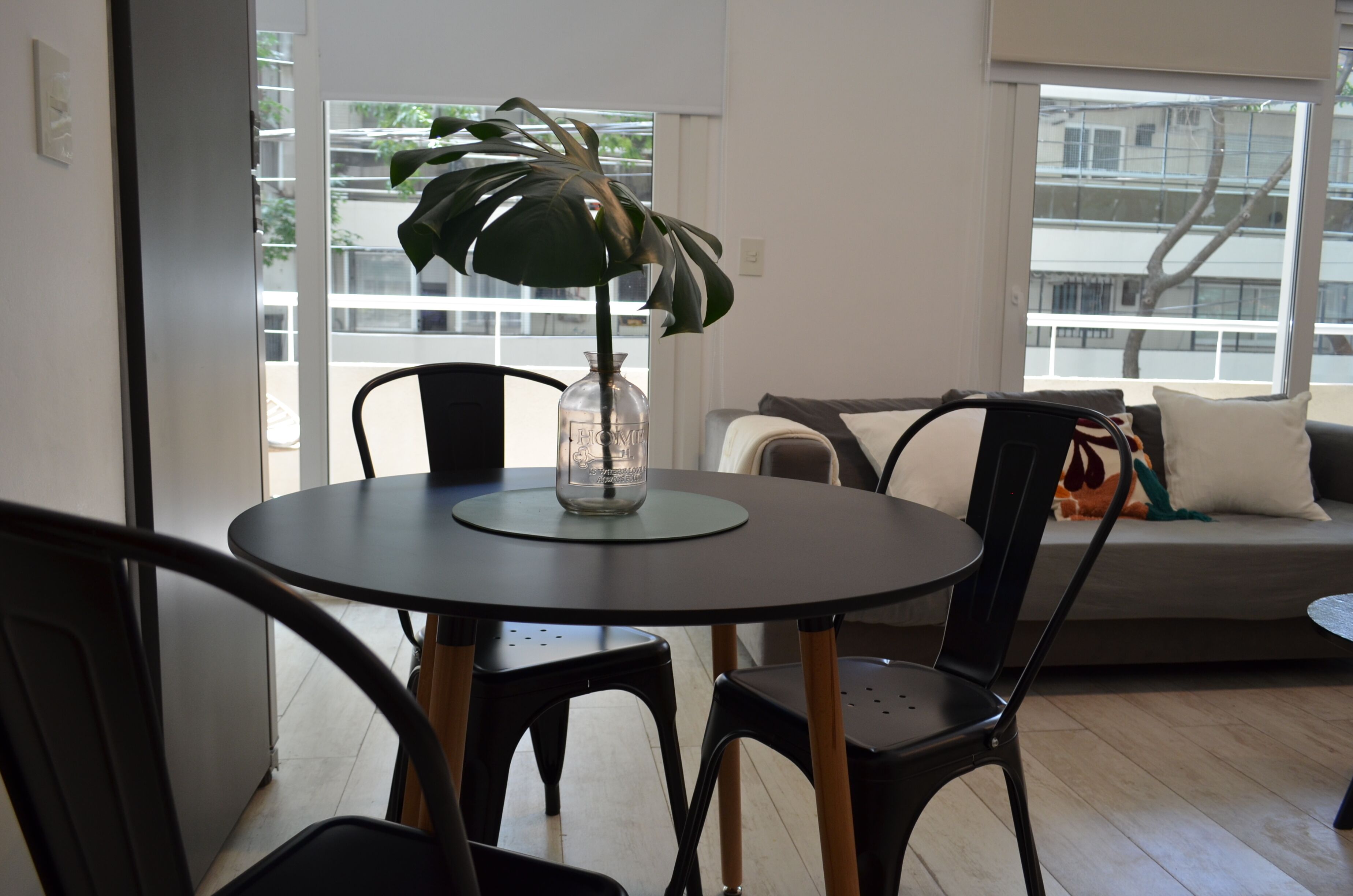 Een moderne woonkamer met een ronde tafel, zwarte stoelen, een bank met kussens en een groot raam met uitzicht op gebouwen en bomen.