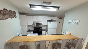Una cocina compacta con armarios blancos, encimera de madera y pileta de acero inoxidable. Hay tres taburetes de bar y un cartel que dice "WELCOME TO OUR HAPPY PLACE".