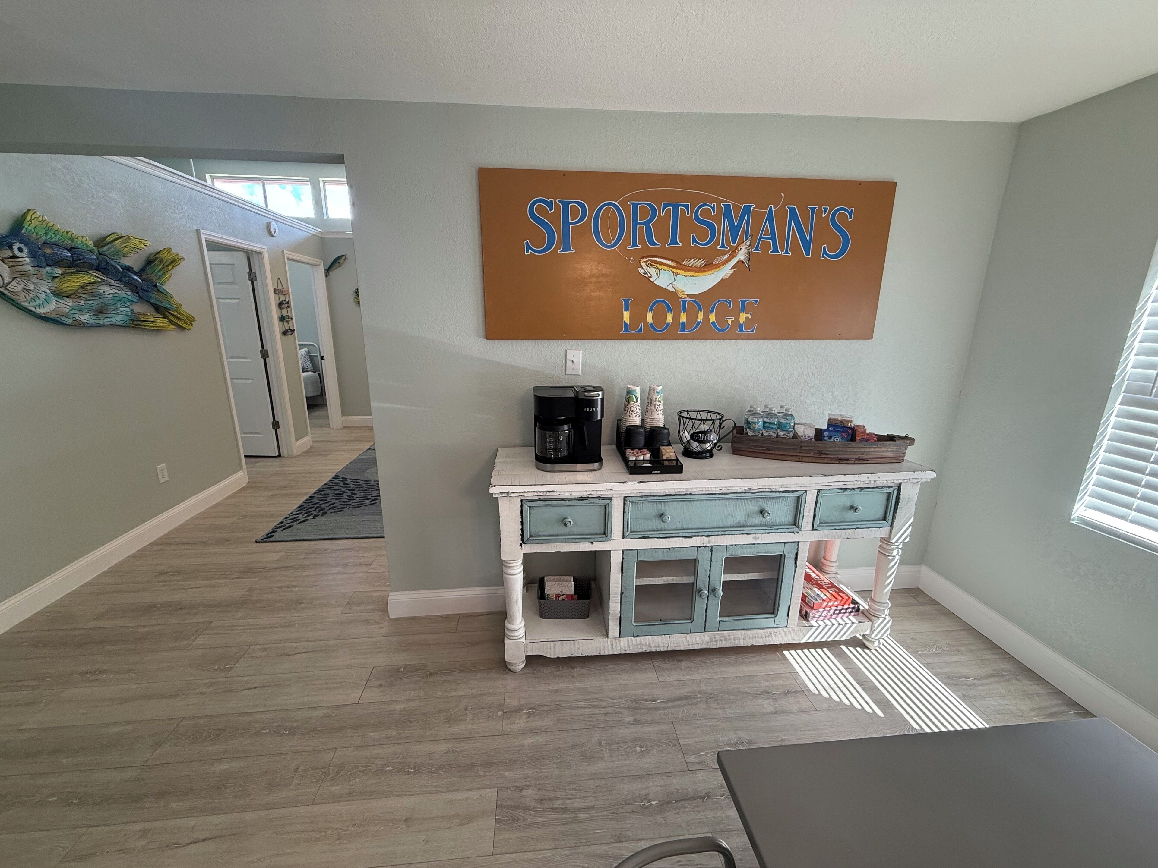A room with a coffee station, a sign reading "SPORTSMAN'S LODGE", and a hallway leading to another room.