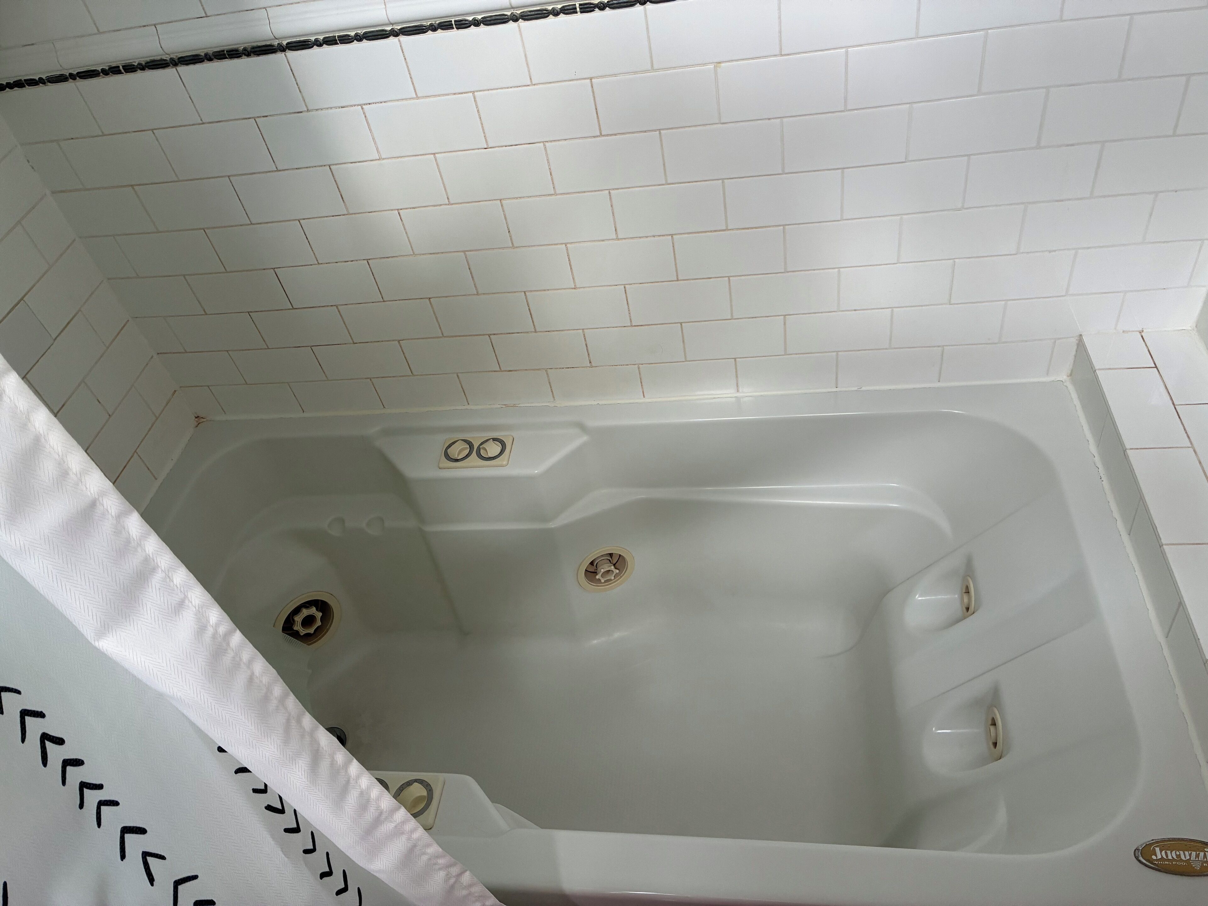 Una bañera blanca con cortina de ducha, paredes de azulejos blancos y desagüe visible.