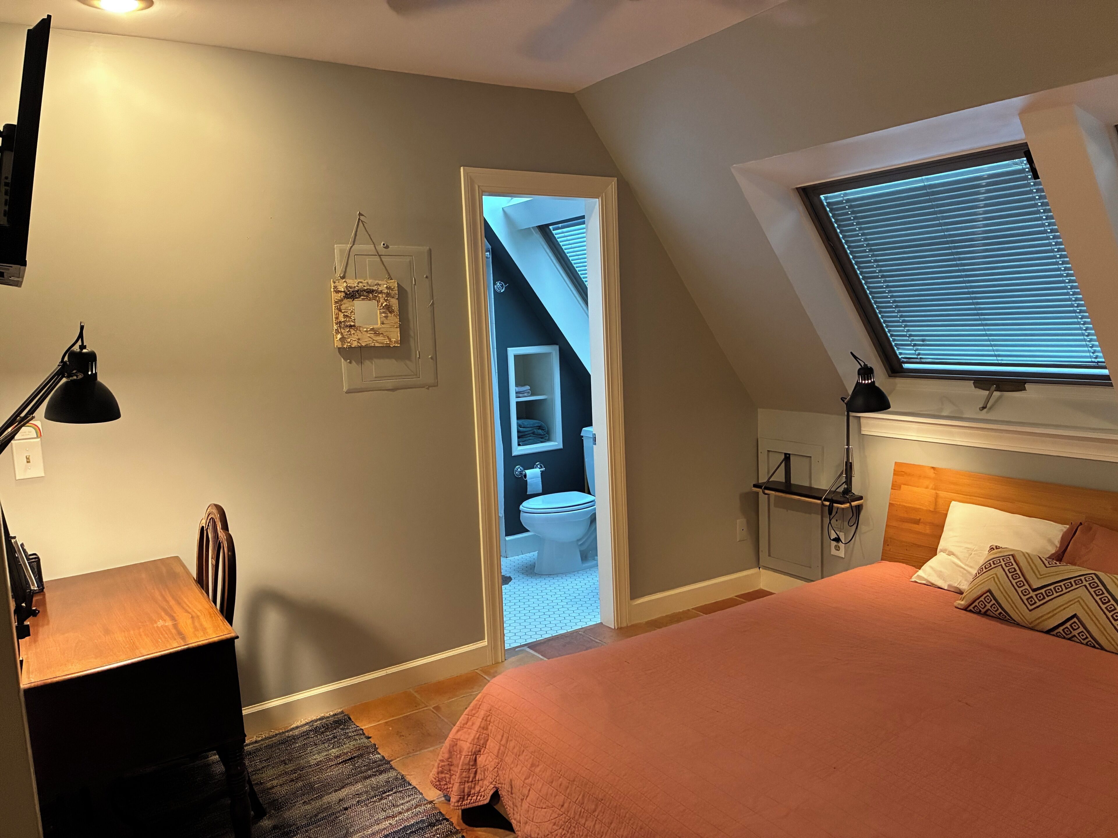 Quarto com cama, escrivaninha, luminária, janela com persianas e porta para um banheiro.