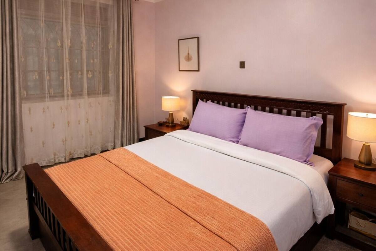 A neatly made bed with a wooden frame, a white and orange bedspread, and two purple pillows. There is a nightstand with a lamp on each side of the bed. The room has a white ceiling with a recessed light fixture.