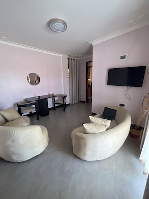 A modern living room with a sofa, a dining table, and a TV mounted on the wall. - The Cozy Home Apartment (Fort Portal)