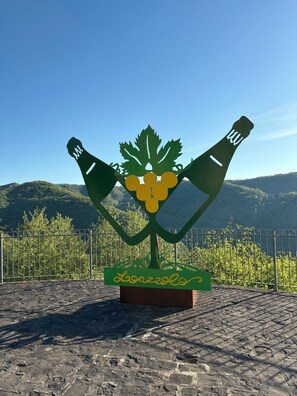 A metal sculpture of a wine bottle and grapes with the word "Lozarolo" on a stone base, set against a mountainous backdrop. - Luce e Forme - Loazzolo (Loazzolo)