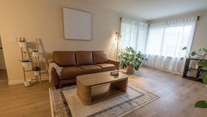 A living room with a brown leather sofa, a wooden coffee table, and a floor lamp. There is a plant on a wooden shelf and another on the floor. - Spacious 7BR 4BA Home - Game Room - AC/Heat - 20 min to Airport (Kent)