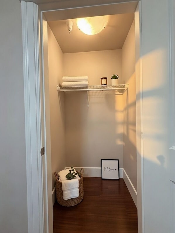 Un pasillo estrecho con suelo de madera, una puerta blanca a la izquierda y una pared blanca. Se observa una pequeña estantería con una vela y una planta, y un letrero de bienvenida enmarcado a la derecha.