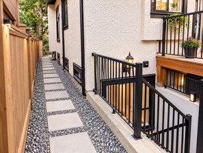 A narrow pathway with stepping stones leading to a building with a wooden fence on one side and a black metal railing on the other.