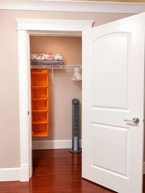 Una habitación con piso de madera, una puerta blanca, un armario abierto con estantes y prendas colgadas, y un calentador portátil.