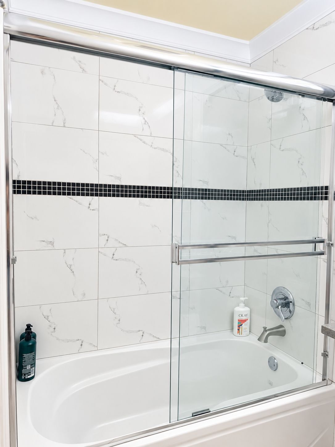 A glass-enclosed shower stall with a white bathtub, marble-patterned walls, and a black tile accent strip.