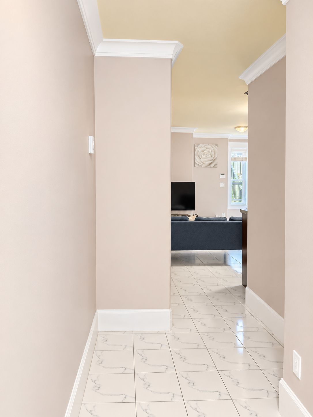 Un couloir au sol en marbre mène à un salon où se trouve un canapé de couleur sombre et un téléviseur à écran plat fixé au mur.
