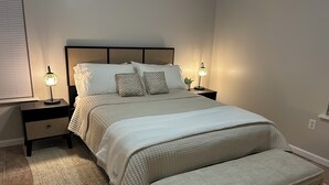 Un dormitorio ordenado con una cama grande, dos mesitas de noche con lámparas, un banco y una ventana con persianas.
