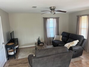 A living room with a black leather sofa, a wooden coffee table, a TV on a stand, and a ceiling fan.