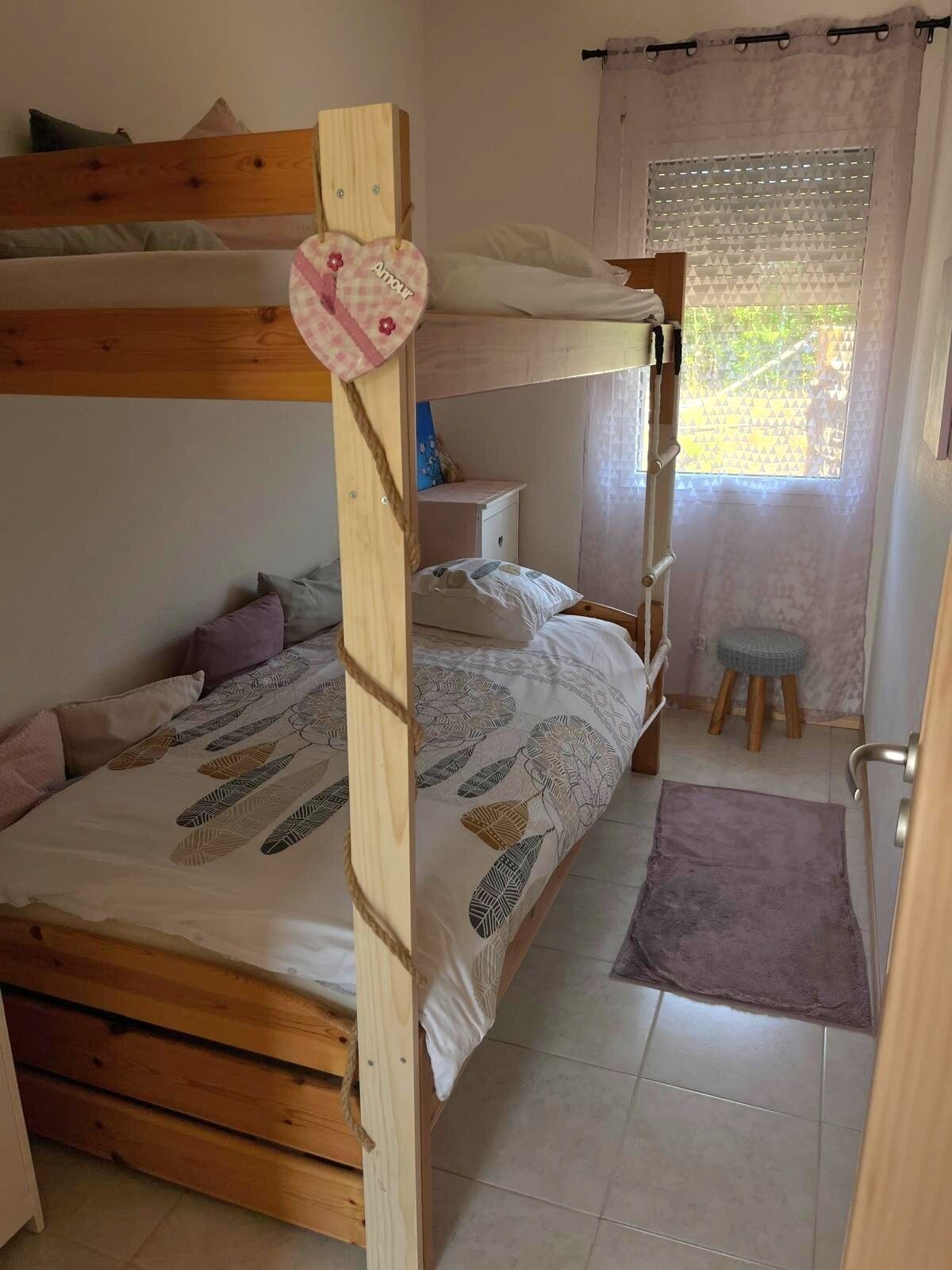A bunk bed with a desk and chair, a small stool, and a window with blinds.
