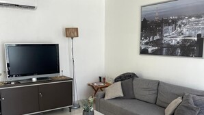 Moderni olohuone, jossa on harmaa sohva, taso-tv jalustalla, pyöreä sohvapöytä, jossa on kasvi, ja kaupunkimaisemaa esittävä taulu seinällä.