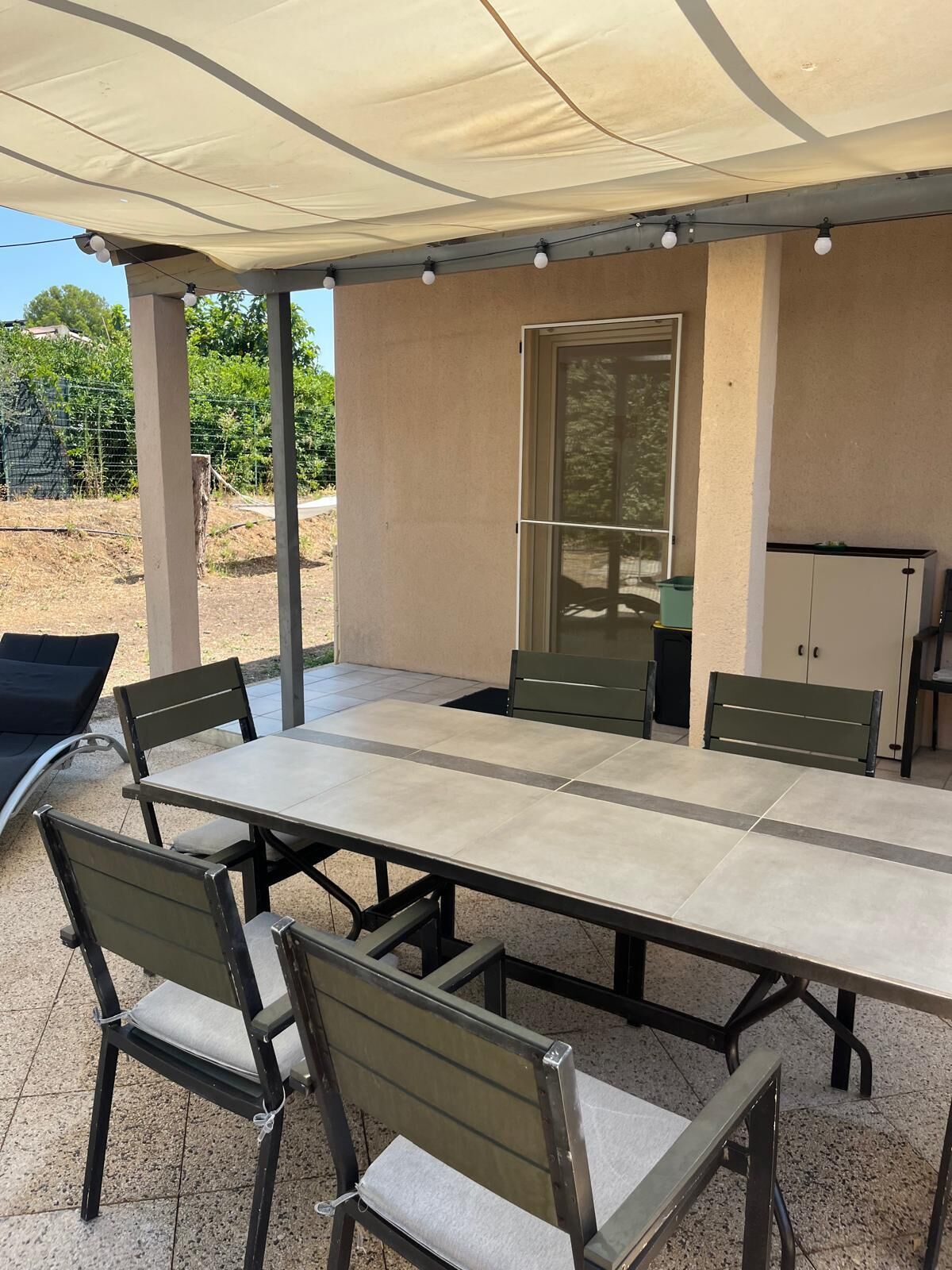 Een terras buiten met een lange tafel, stoelen, een luifel en een raam.