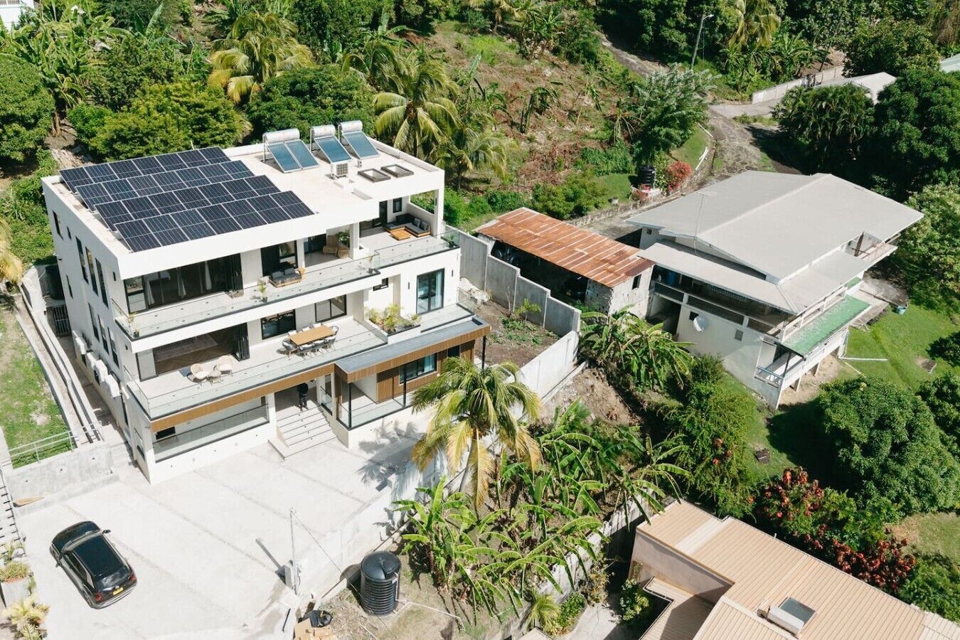 Une maison moderne à deux étages, dotée de panneaux solaires sur le toit, avec une voiture garée dans l’allée et une végétation luxuriante qui entoure la propriété.