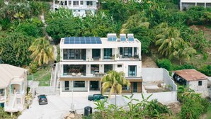Un bâtiment moderne à deux étages, doté de panneaux solaires, entouré d’une végétation luxuriante et de palmiers.