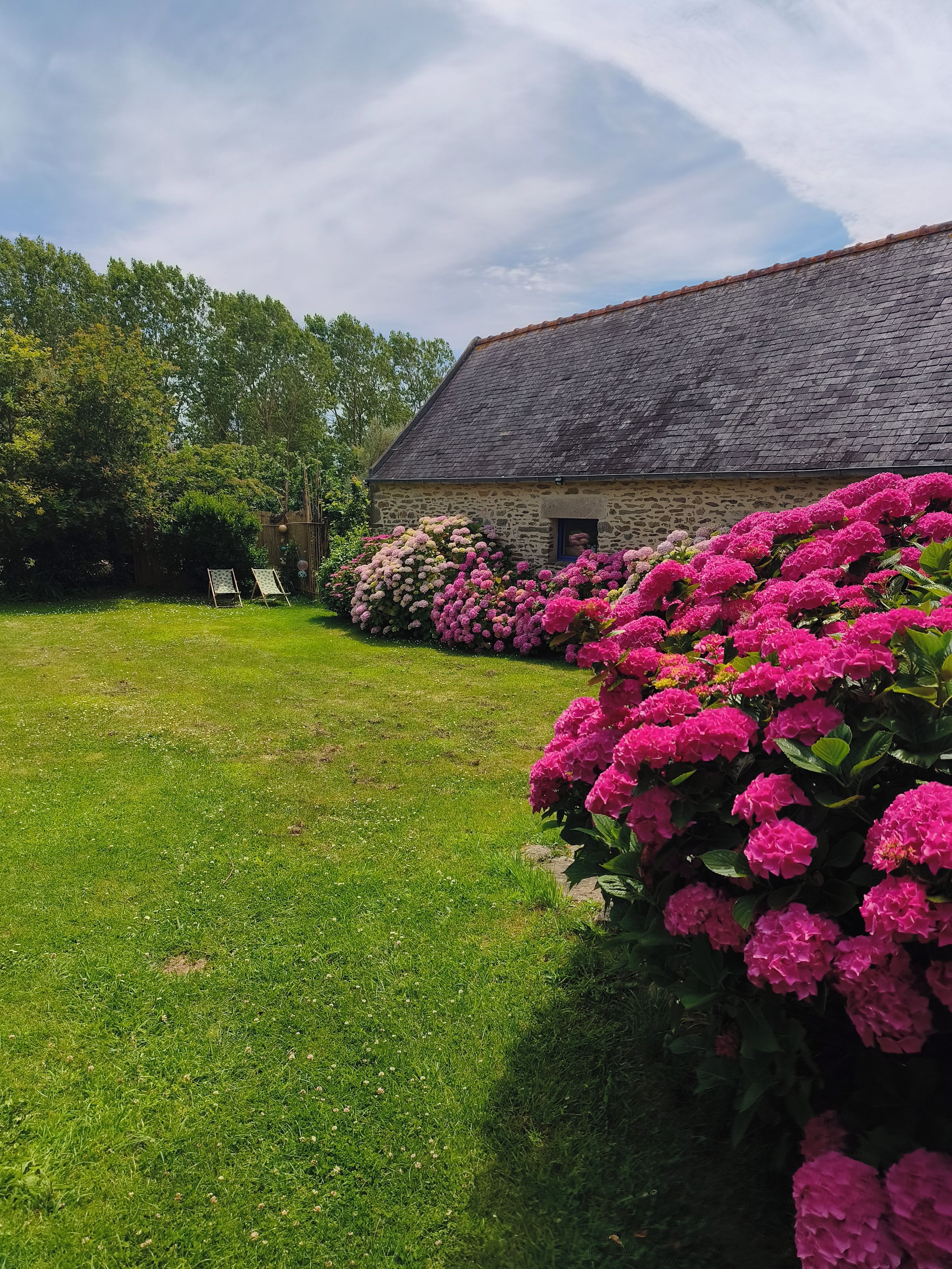 A stone building with a sloped roof, surrounded by lush green grass and vibrant pink flowers.