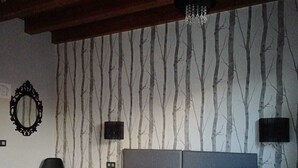 Een tweepersoonsbed met witte lakens, een grijs hoofdbord en twee nachtkastjes met lampen. Een spiegel hangt aan de muur, en er staat een stoel in de hoek.