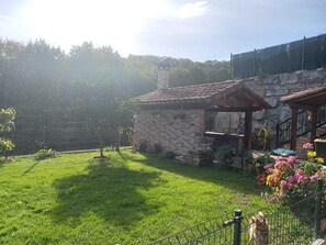 Un bâtiment en pierre avec un toit en tuiles, un petit jardin avec de la pelouse et des fleurs, et une clôture en métal.