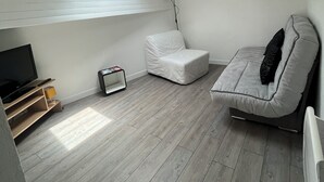 Una habitación pequeña y moderna con techo inclinado, un sofá blanco, una pequeña silla blanca, un televisor y un radiador.