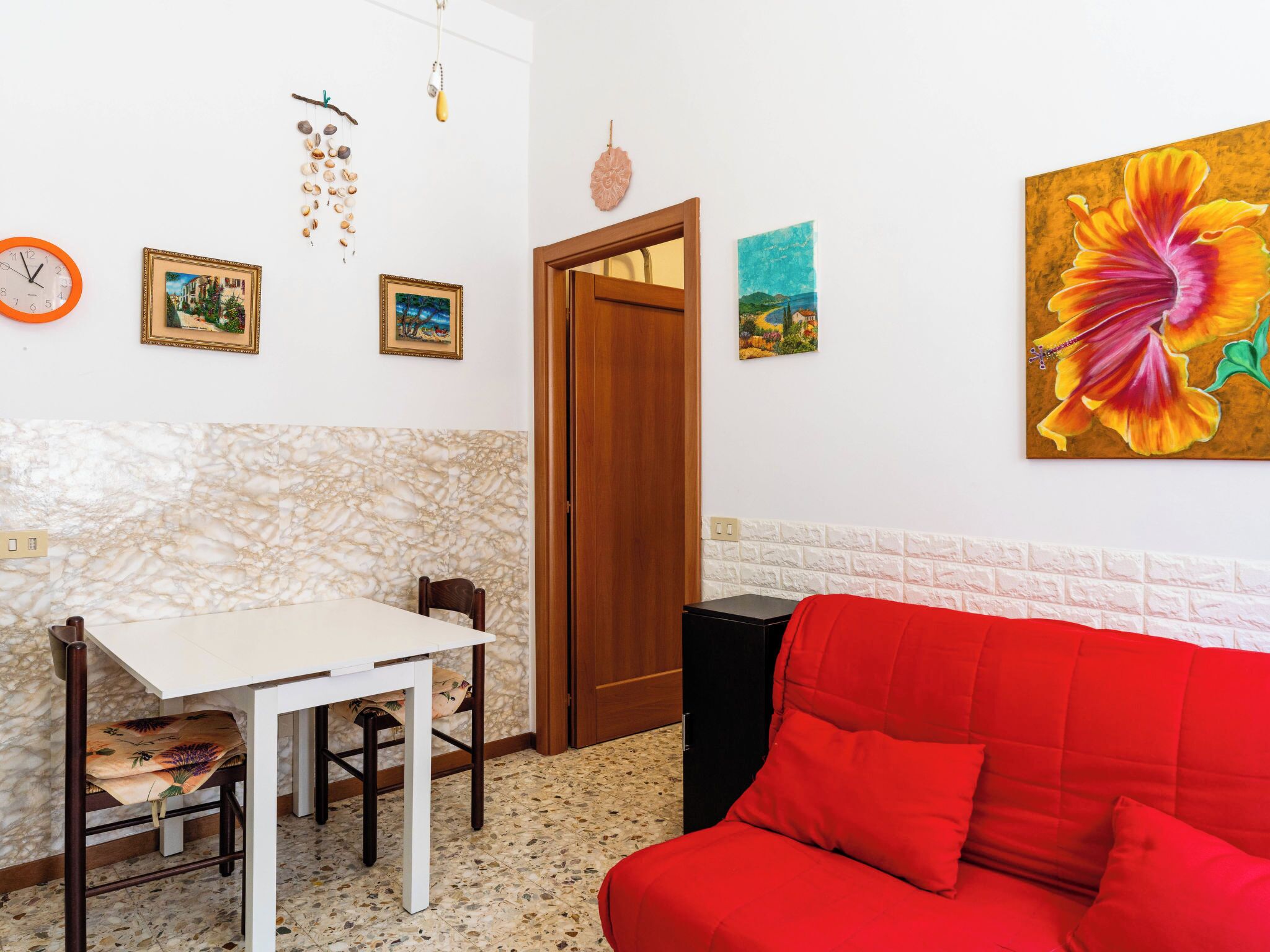 A room with a dining table, chairs, a red sofa, a wooden door, and wall art including a clock, framed pictures, and a painting of a flower.
