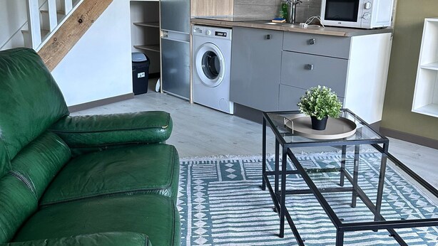 Een moderne keuken met een wasmachine, magnetron en een zithoek met een groene bank en een glazen tafel.