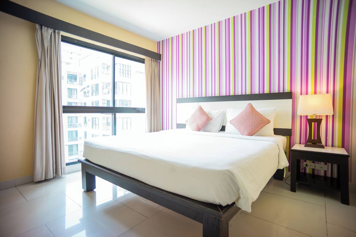 A hotel room with a large bed, a nightstand with a lamp, a window with curtains, and a striped wall.