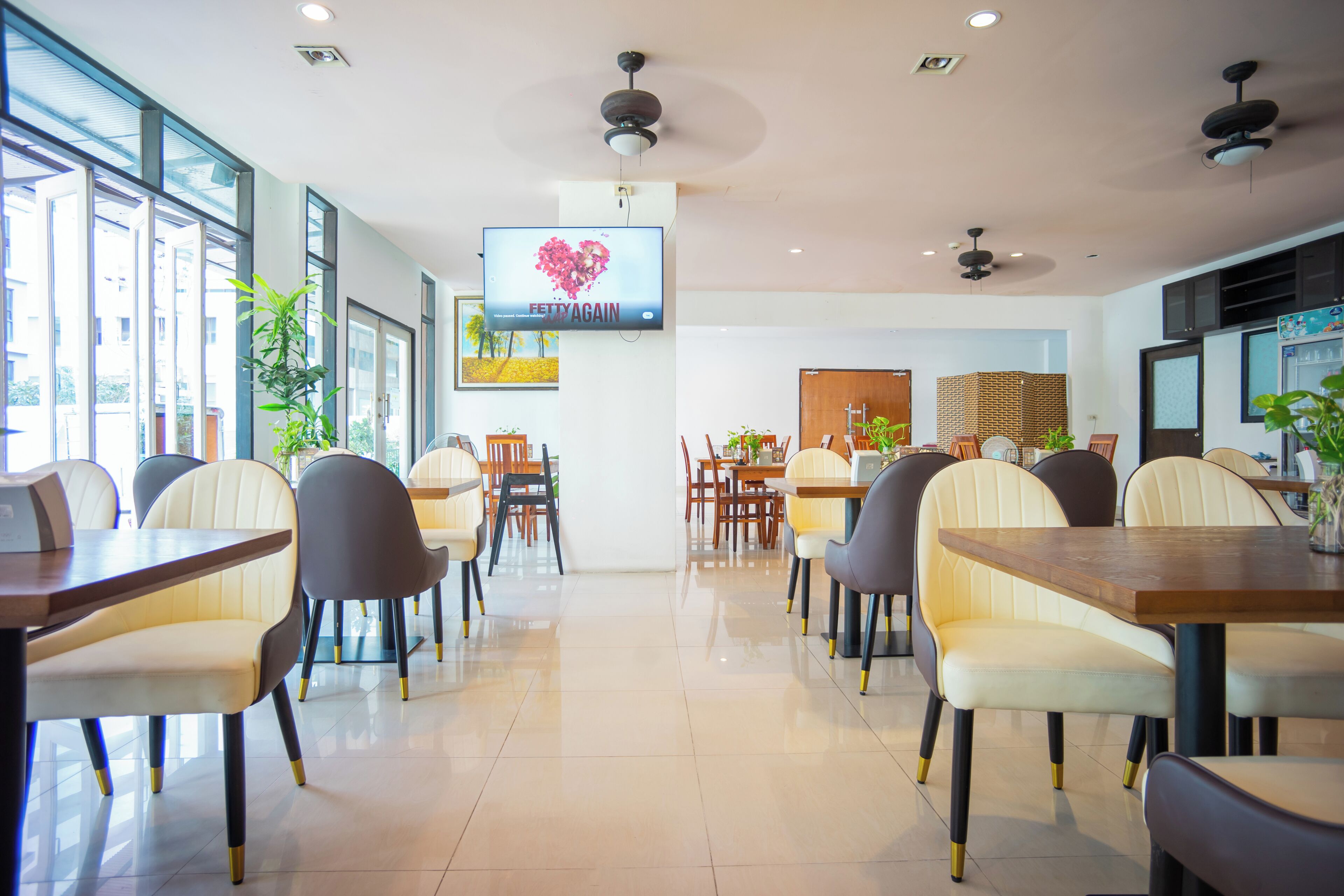 A modern dining area with multiple tables and chairs, a television displaying a heart-shaped image, and large windows allowing natural light.
