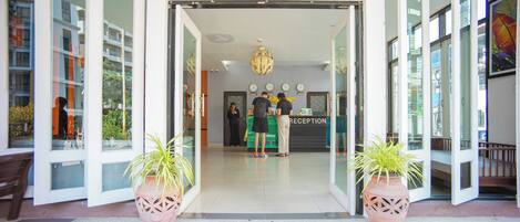 A hotel entrance with a reception desk, a 'WELCOME' mat, and potted plants.