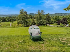 Um campo de golfe com uma cadeira em formato de bola de golfe grande, com a placa 'The Woods' em primeiro plano.