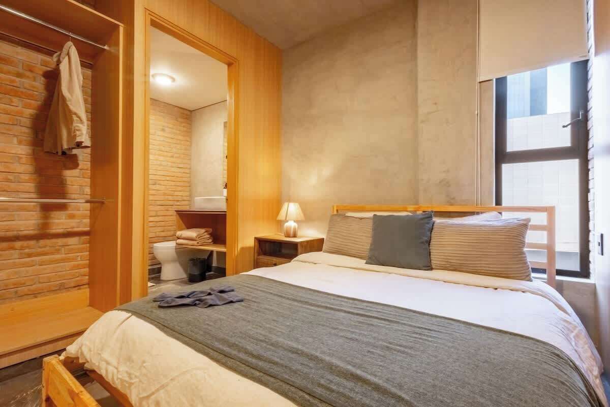 Une chambre avec un lit en bois, une table de chevet avec une lampe, un mur en briques et une fenêtre.